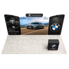 Full-width front perspective capturing the complete 20ft exhibit footprint with illuminated graphics, overhead branding, angled wing panels, and podium positioned at the front.